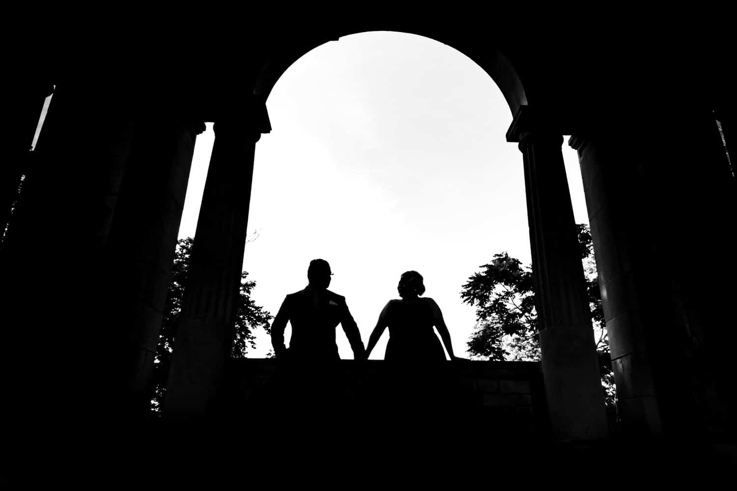 A dramatic black and white silhouette of a man and woman standing side by side underneath an arch. 