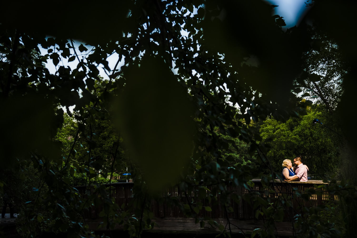 A colorful picture taken through the leaves of a tree of a man and woman standing on a bridge, leaning in to share a kiss during their Loose Park engagement session in Kansas City. 