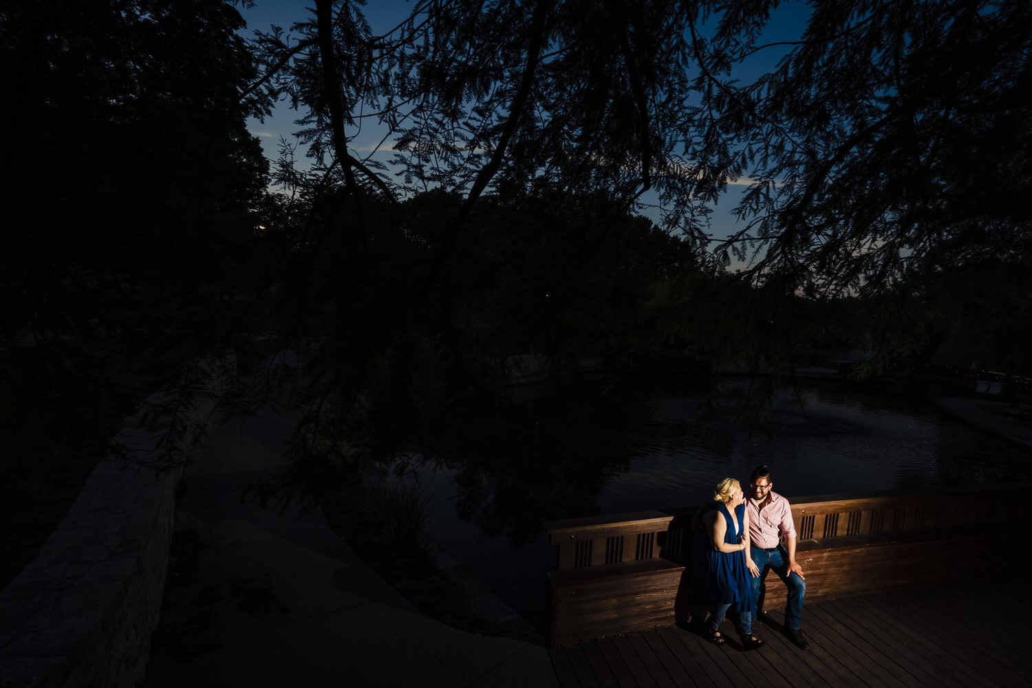 A wide picture of a man and woman sitting on a bridge, the sunset visible behind them during their romantic engagement session at Kansas City's Loose Park. 