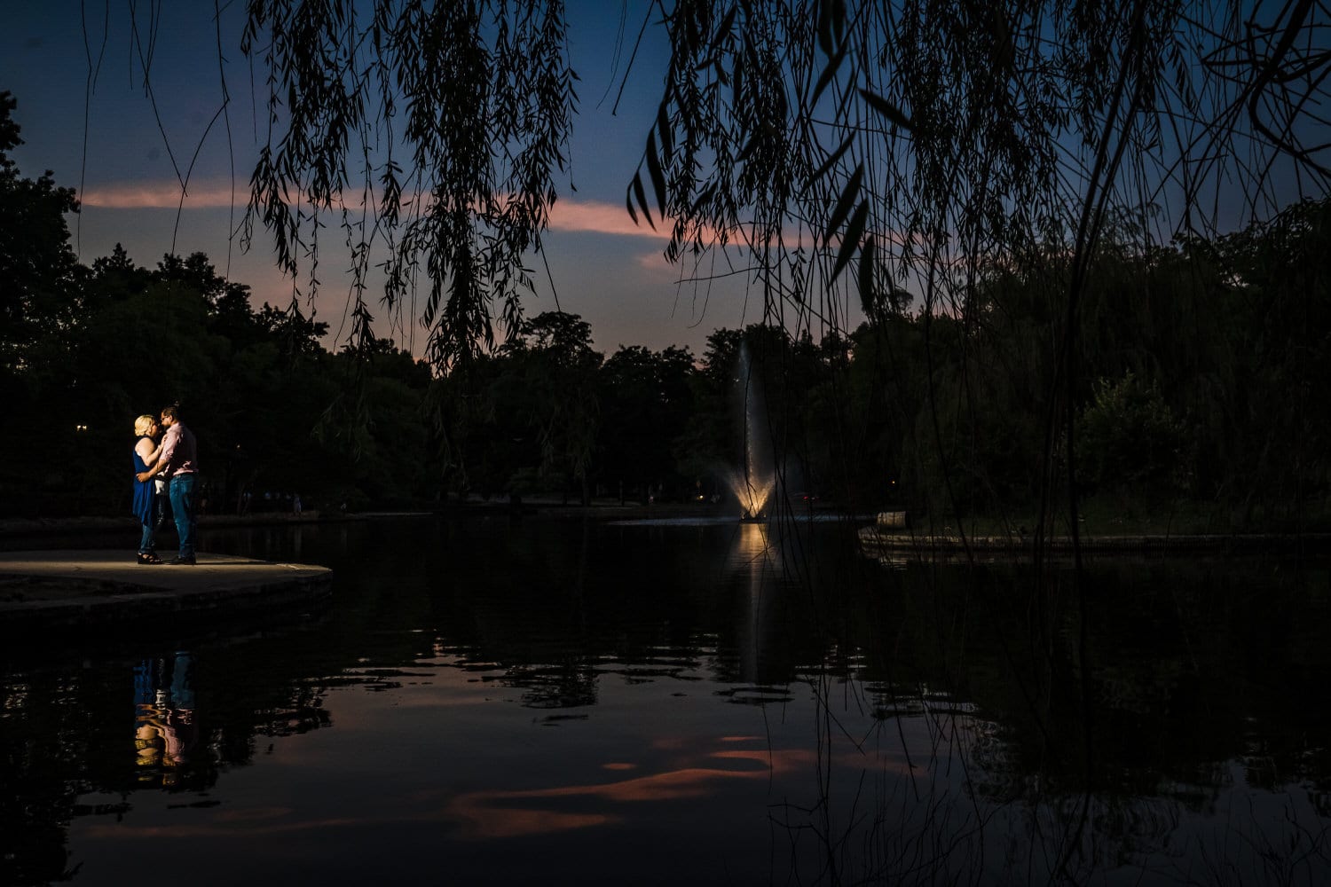 A dramatic portrait of a man and woman standing next to the pond at Loose Park, the fountain lit up behind them in front of the sunset during their engagement session. 