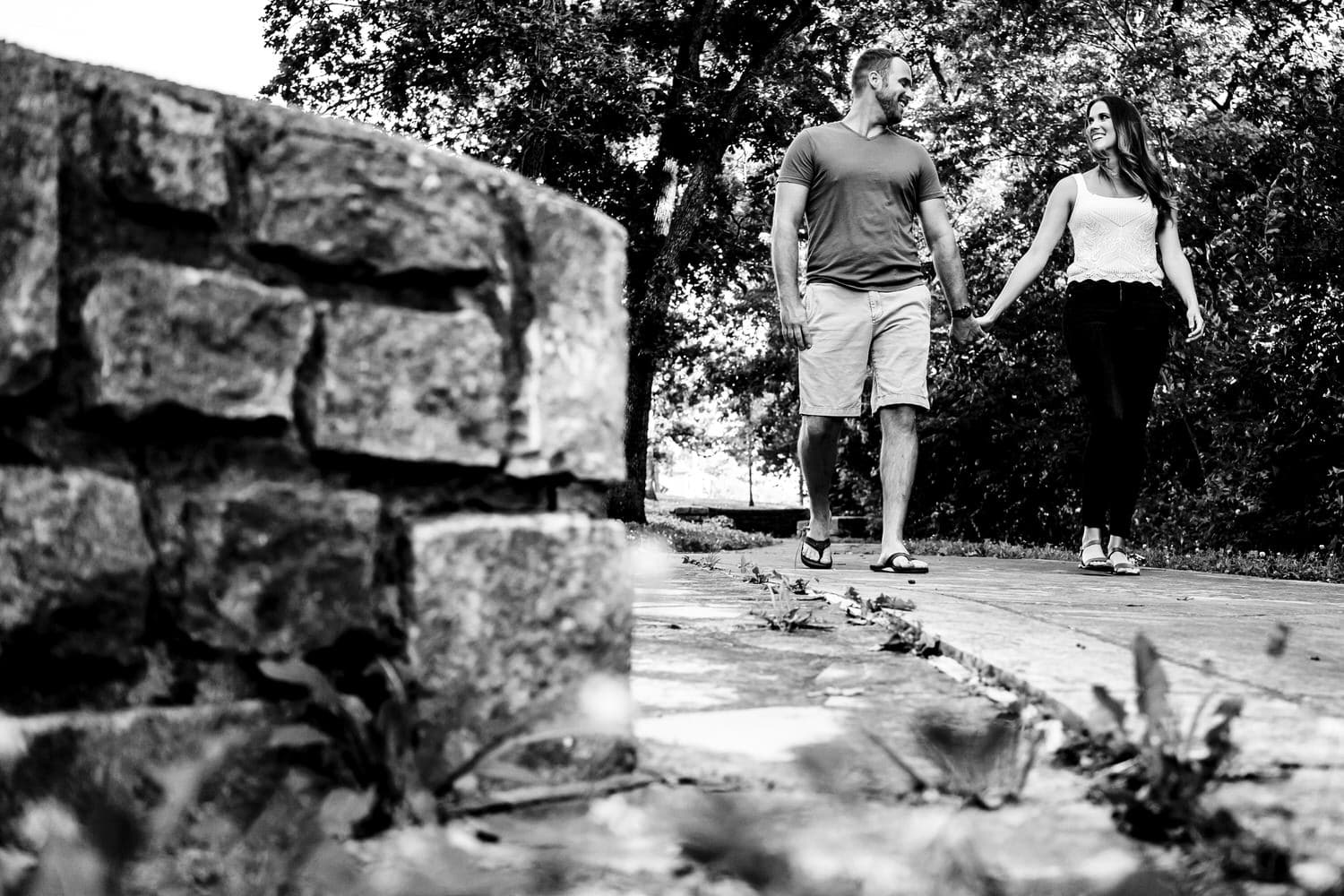 A candid black and white picture of an engaged couple holding hands walking down a path during their summer engagement session at Swope Park in Kansas City. 