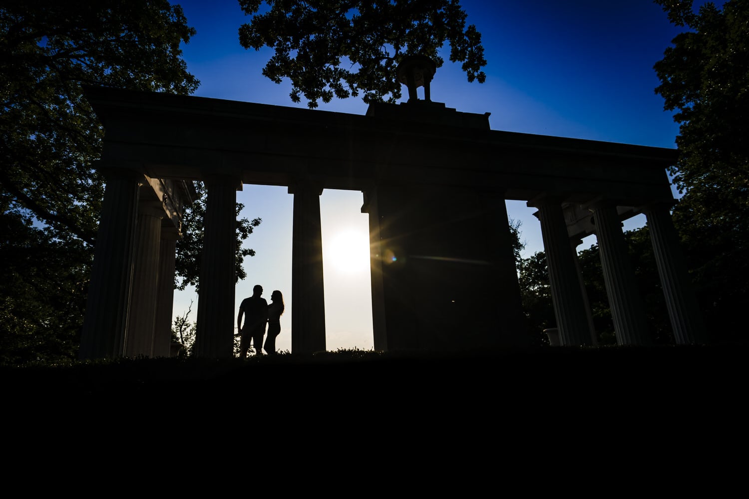 A colorful silhouette of an engaged couple standing between columns, lit from behind during a summer engagement session in Kansas City. 