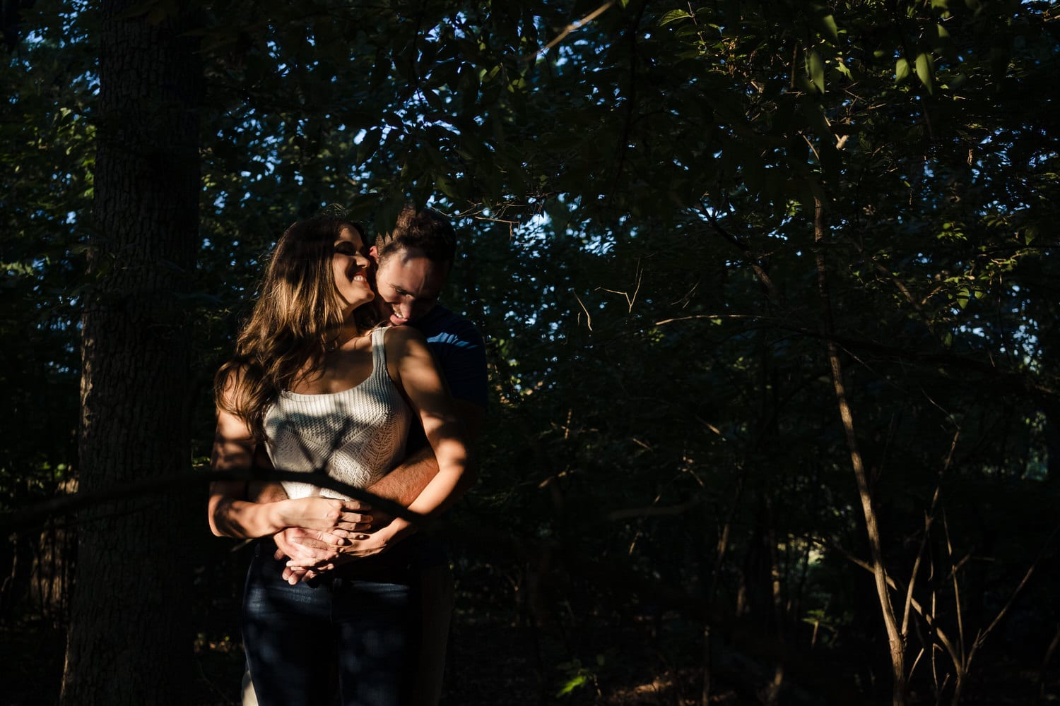 A candid, moody portrait of an engaged couple sharing an embrace surrounded by trees during their summer engagement session in Kansas City. 