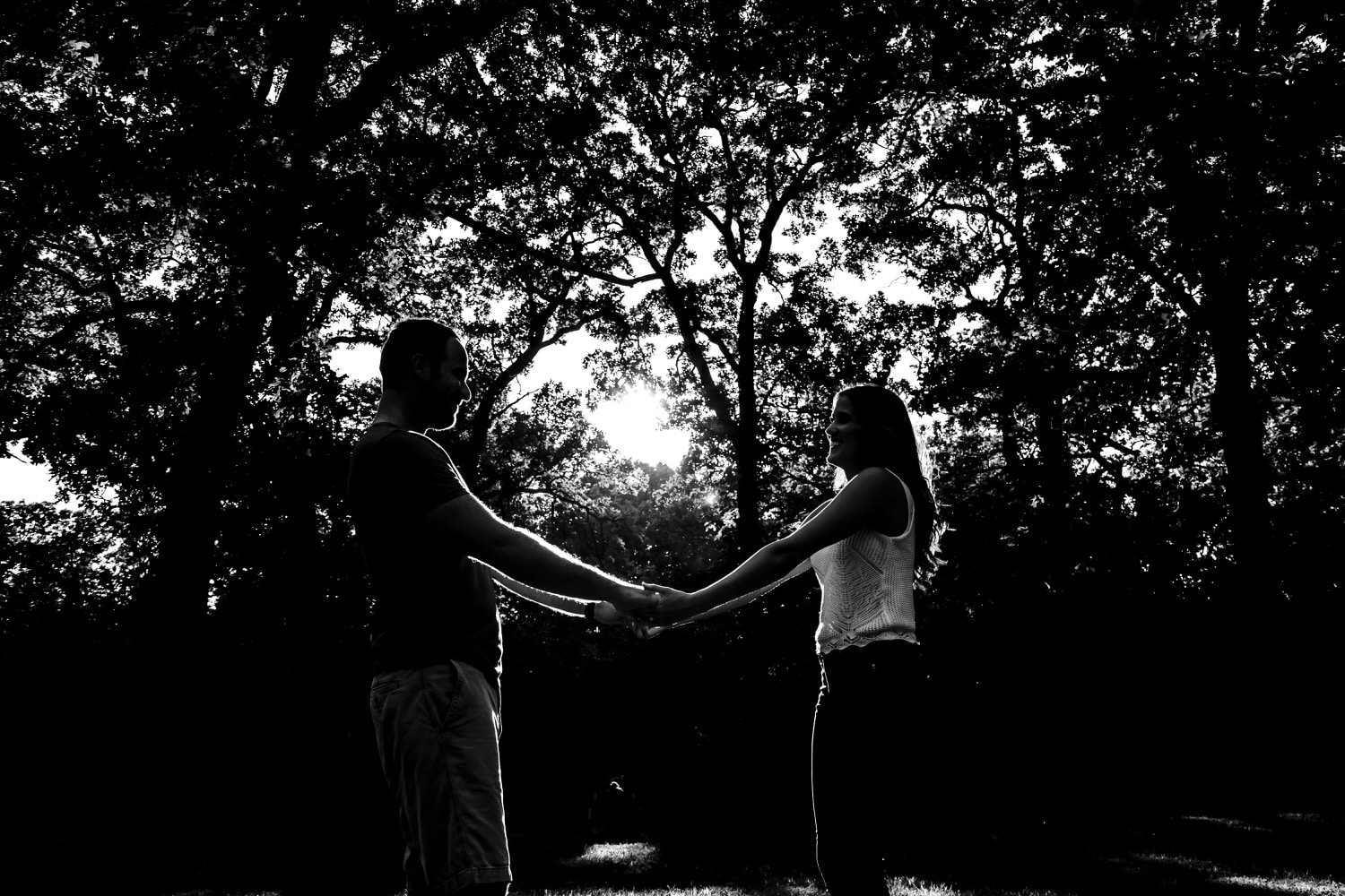 A black and white, wide portrait of an engaged couple holding hands in a wedge of sunlight during their engagement session at Swope Park. 