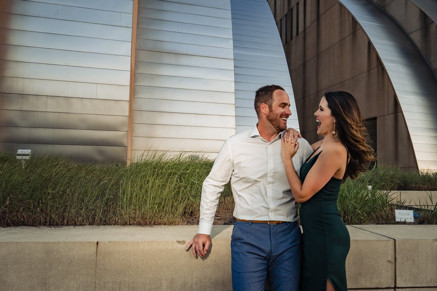 A candid, colorful picture of an engaged couple laughing together as they share an embrace during their summer engagement session at Kauffman Center for the Performing Arts in Kansas City. 