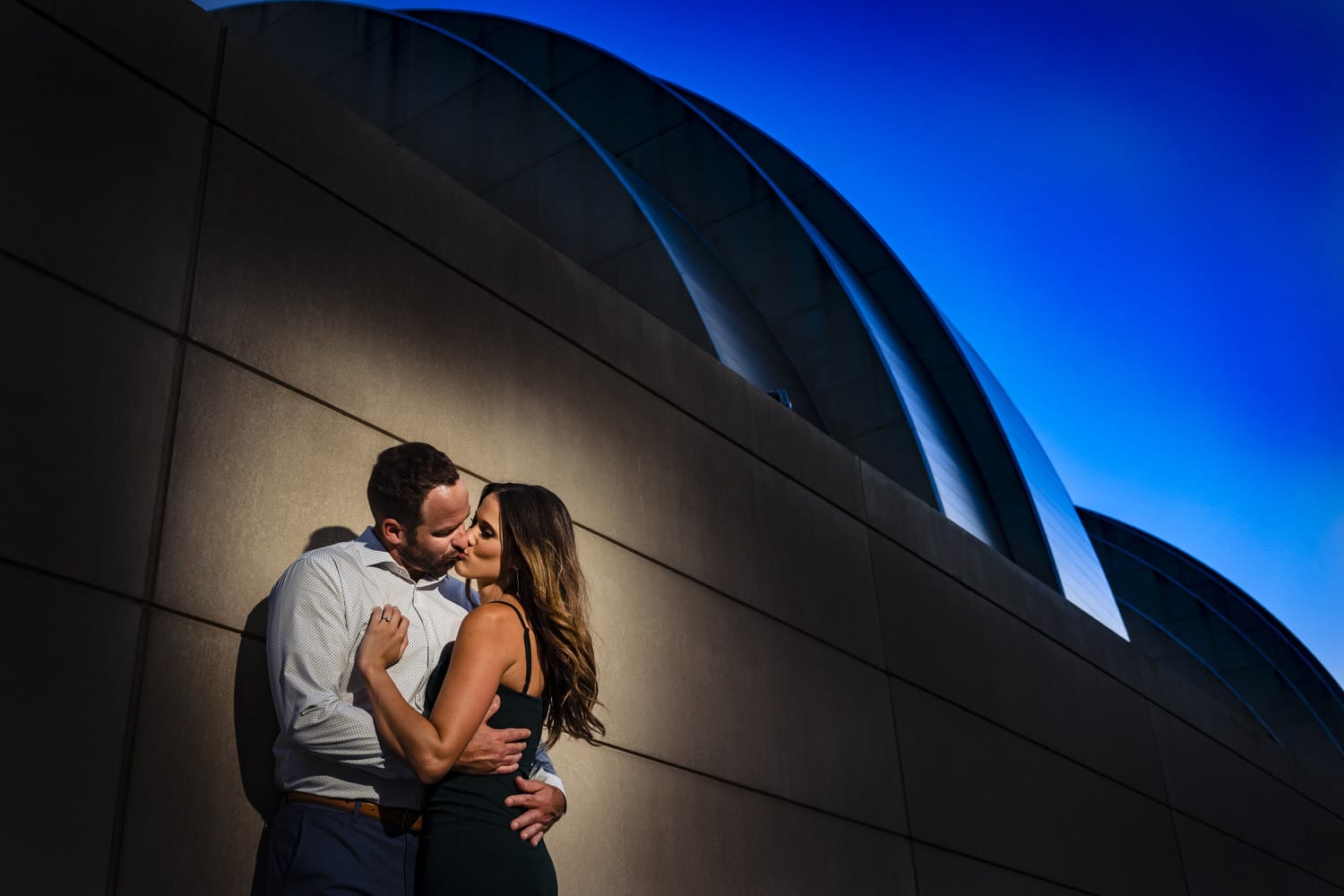 A colorful, candid picture of an engaged couple leaning against the retaining wall of the Kauffman Center for the Performing Arts during their summer engagement session in Kansas City. 