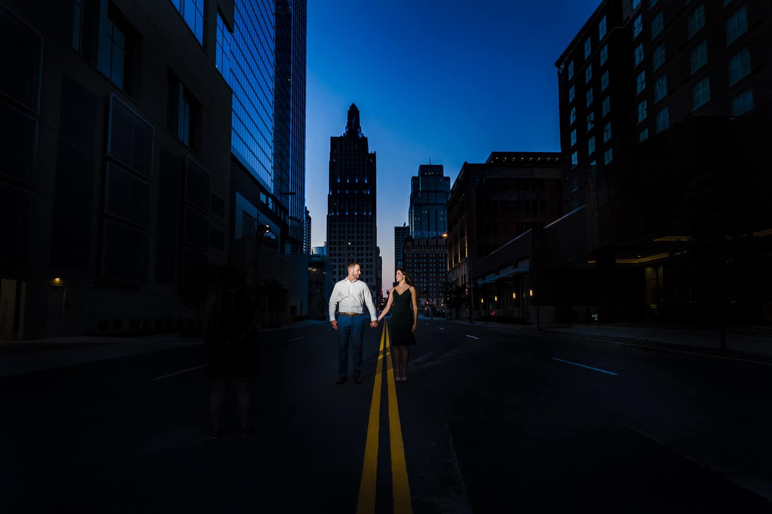 A wide, colorful picture of an engaged couple holding hands as they walk down the center of a downtown Kansas City street during their summer engagement session. 