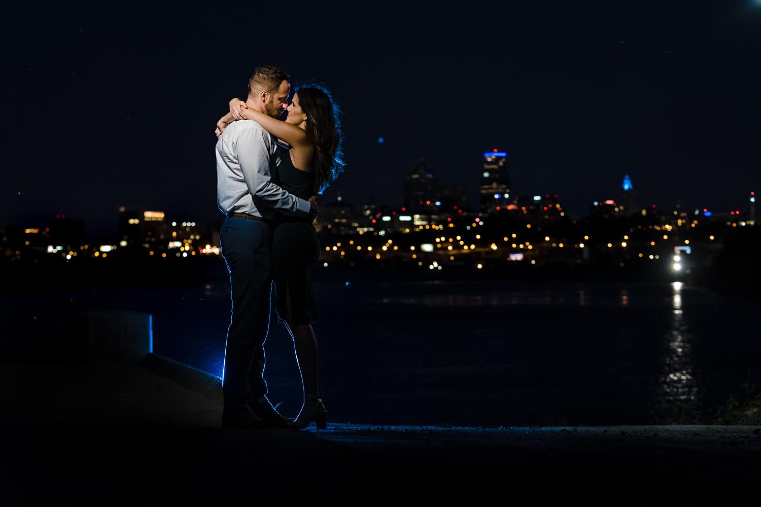 A wide, colorful portrait of an engaged couple sharing an embrace as the Kansas City skyline lights up behind them during their summer engagement session in Kansas City. 