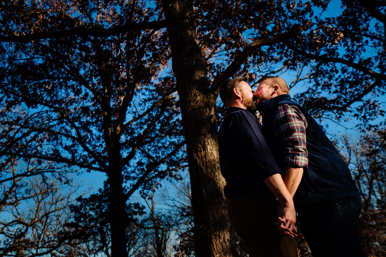 A candid, colorful picture of an engaged couple holding hands and sharing a kiss under a canopy of fall leaves during their fall engagement session in Kansas City. 