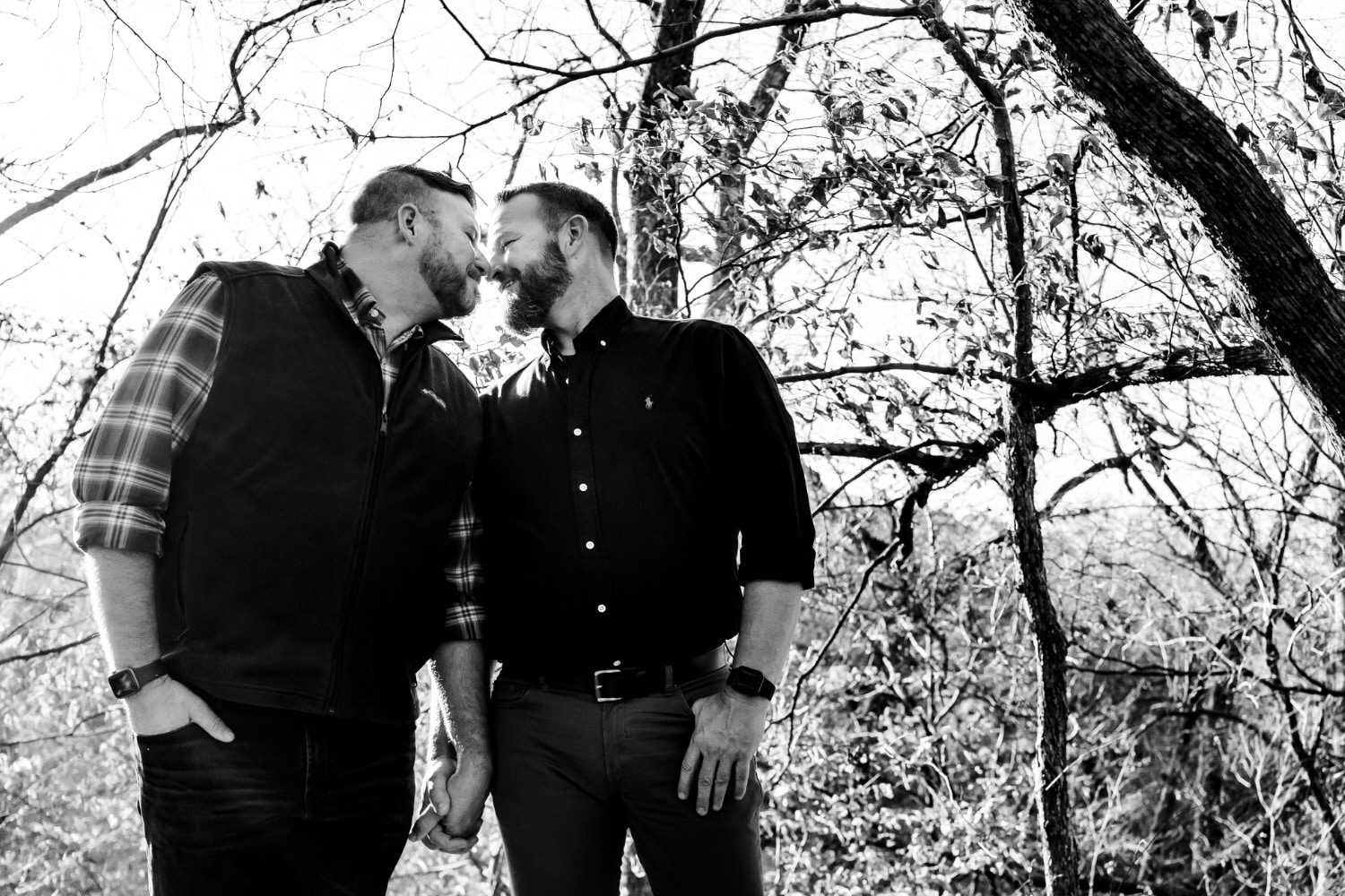 A candid, black and white picture of an engaged couple holding hands and sharing a kiss under a canopy of fall leaves during their fall engagement session in Kansas City. 