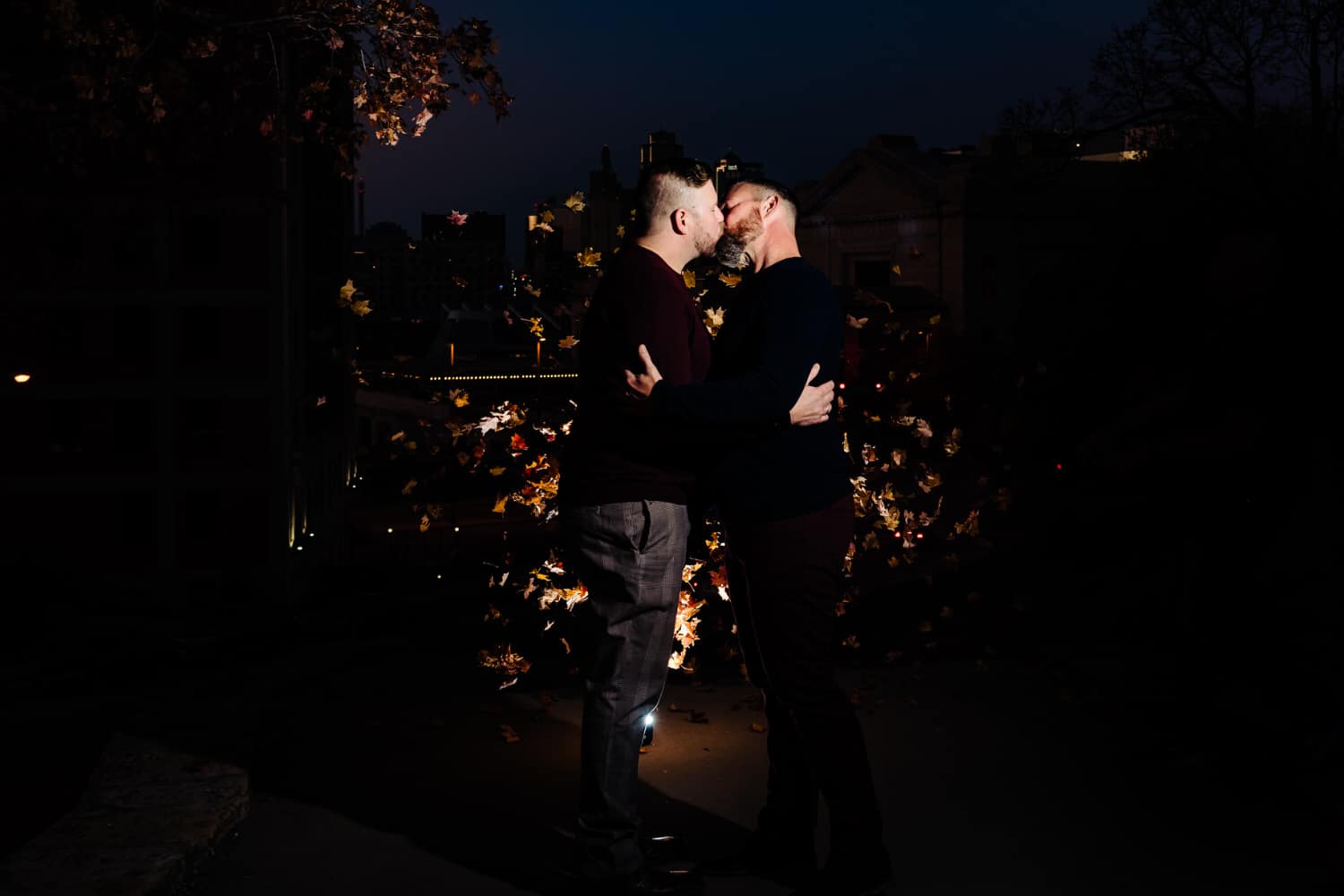 A candid portrait of an engaged couple leaning in to share a kiss as leaves rain down around them during a fall engagement session in Kansas City. 