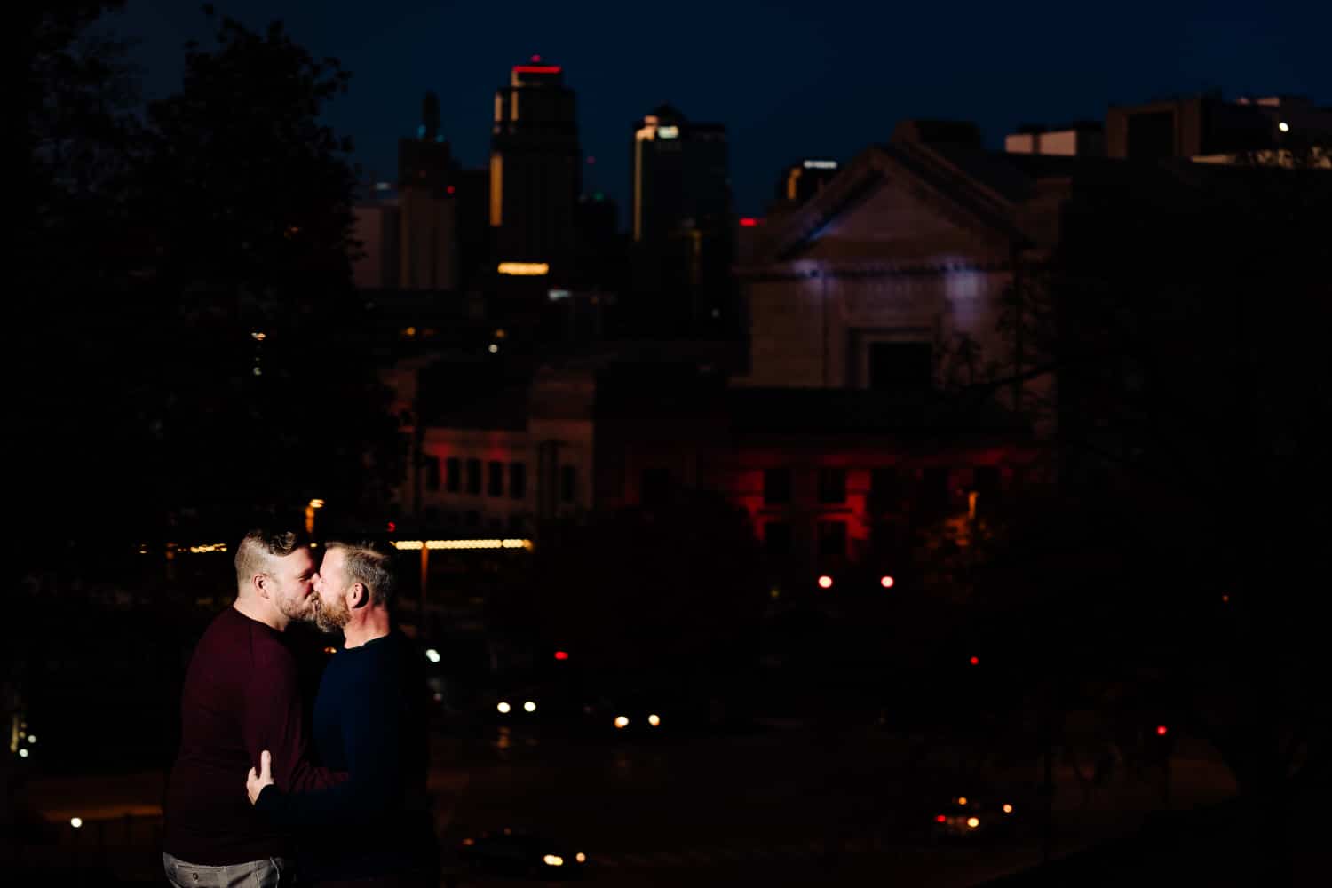A portrait of an engaged couple sharing a kiss during their nighttime engagement session, the Kansas City skyline visible behind them. 