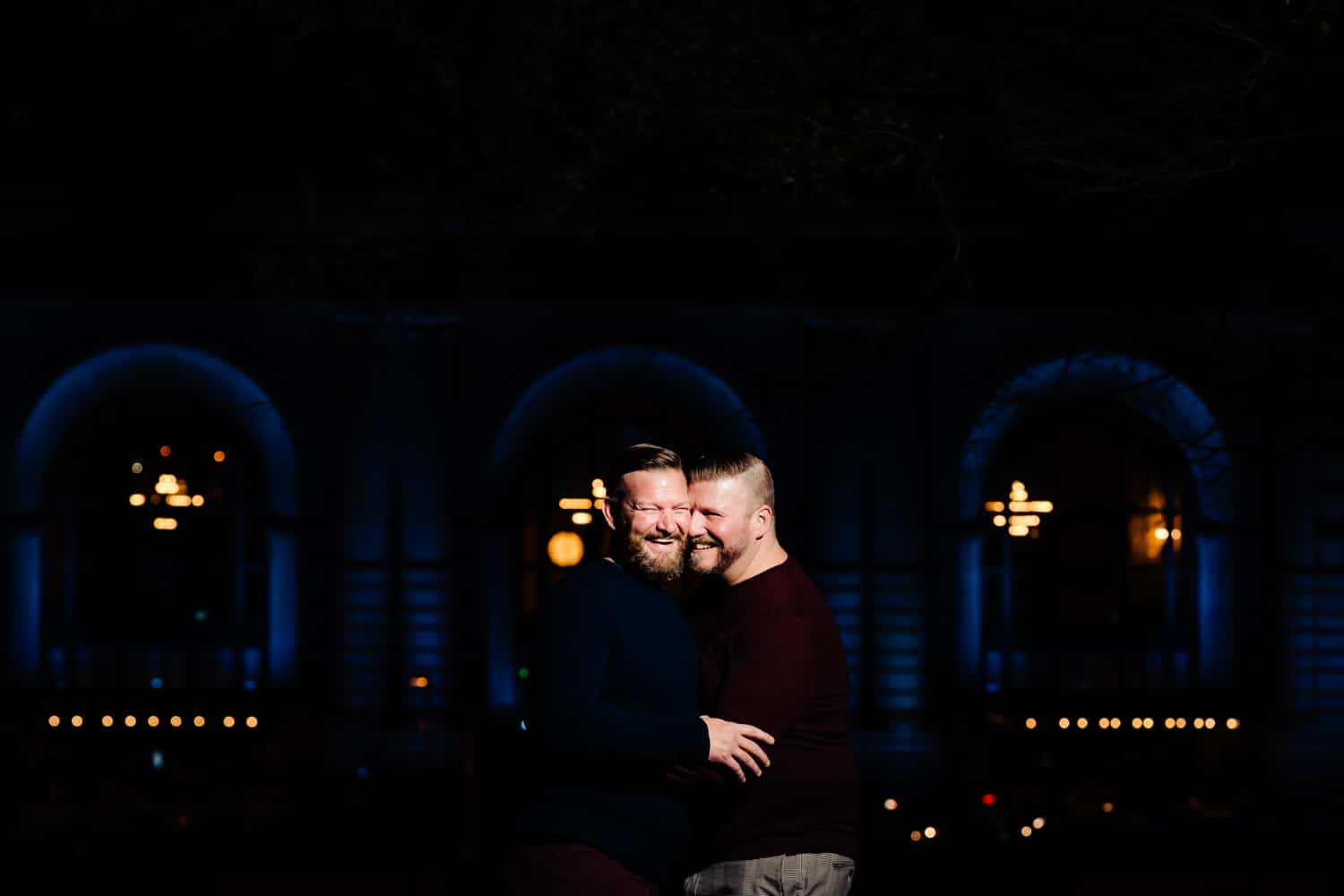 A candid picture of an engaged couple sharing an embrace and laughing together during a nighttime engagement session, Union Station visible in the background. 