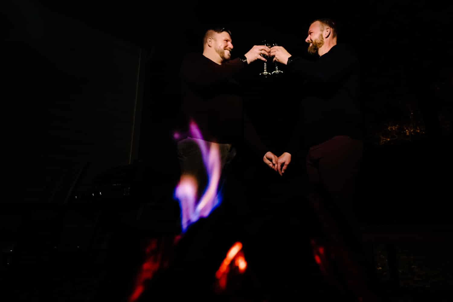 A candid picture of an engaged couple clinking champagne flutes together as they sit by a fire in their backyard during a fall engagement session in Kansas City. 