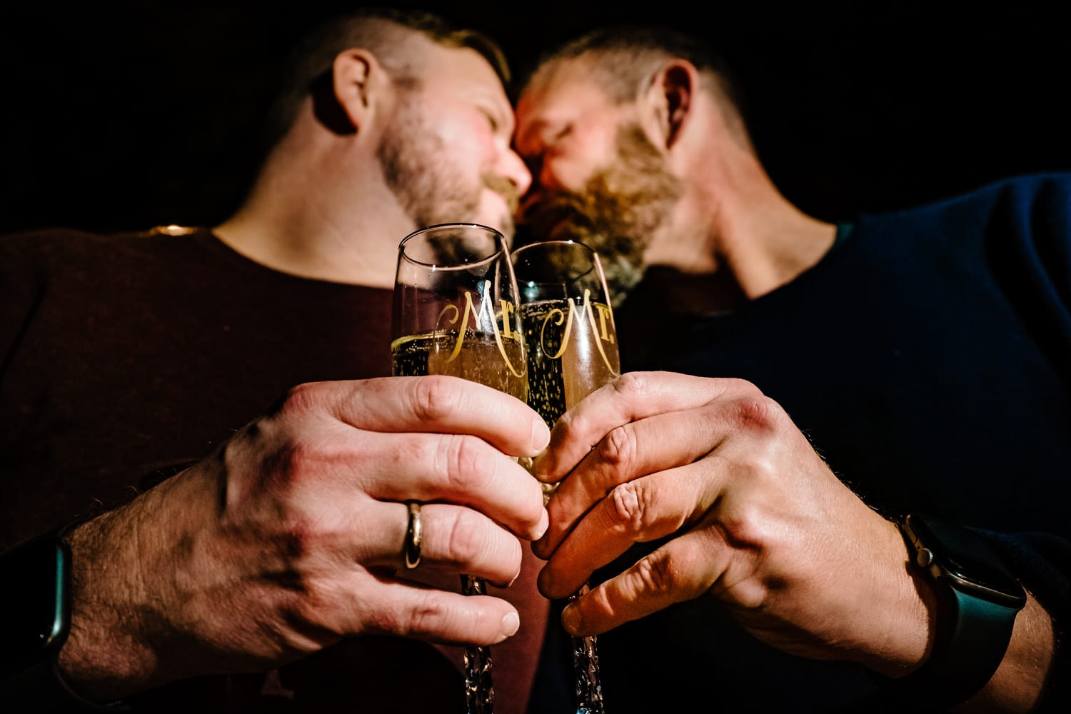 A close-up picture of two champagne flutes that say "Mr." on them, with an engaged couple leaning in towards each other in the background to share a kiss. 