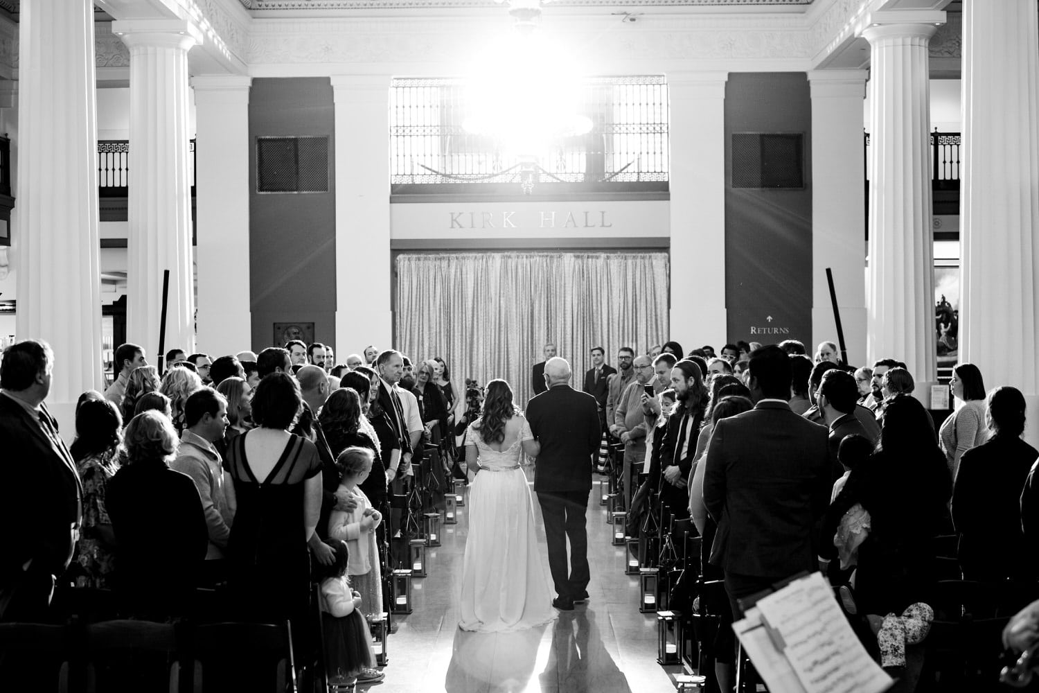 A black and white candid picture of a bride's father escorting her down the aisle during her wedding at The Kansas City Public Library downtown branch. 