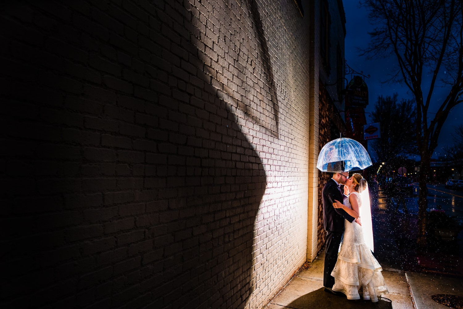 A colorful portrait of a bride and groom sharing a kiss underneath a clear umbrella on their rainy wedding day in Kansas City. 