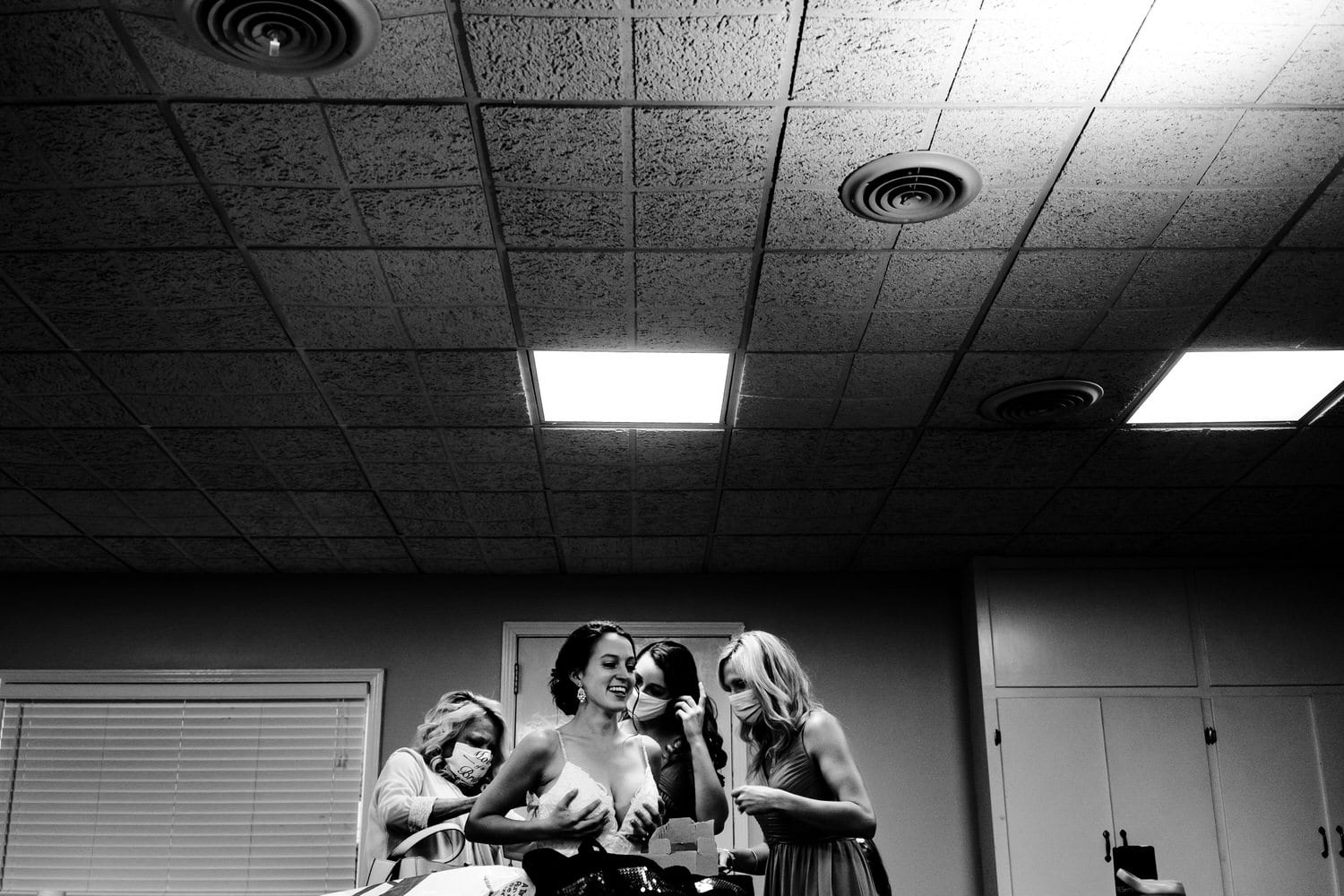 A black and white picture taken from across a table of a bride holding her wedding dress up as her mom and sister lean in to zip it up on the day of an intimate church wedding ceremony in Kansas City. 