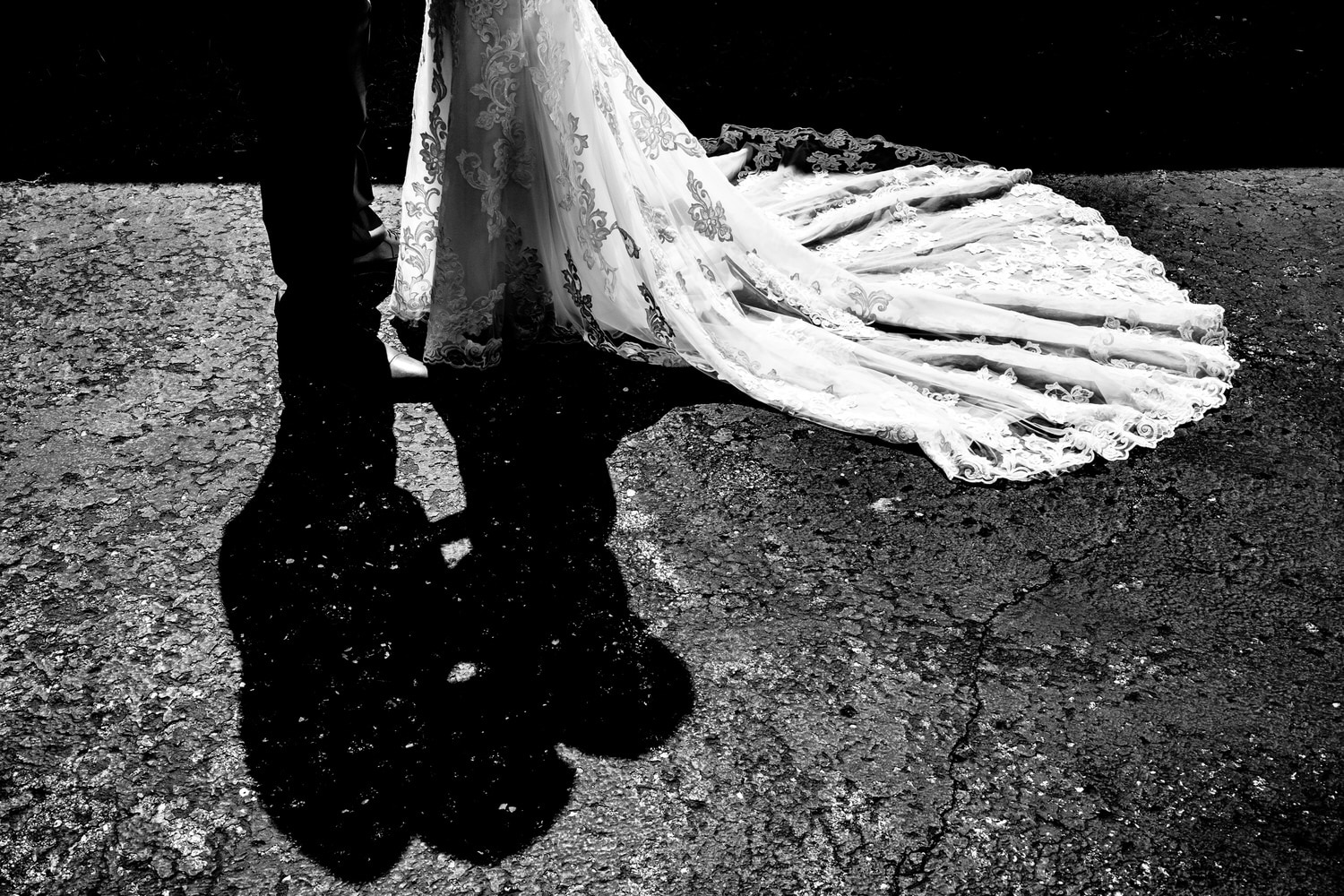A black and white picture of the bottom half of a bride and groom, their top halves visible in shadow form beneath them as they lean in to share a kiss on their wedding day. 