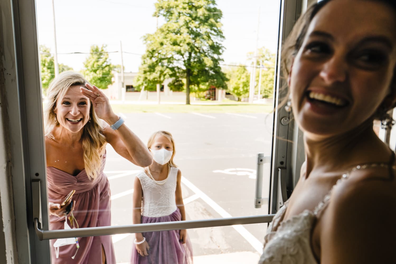 A candid colorful picture of a bride looking over her shoulder at her groom as the bride's sister and niece look through the glass door at the bride. 