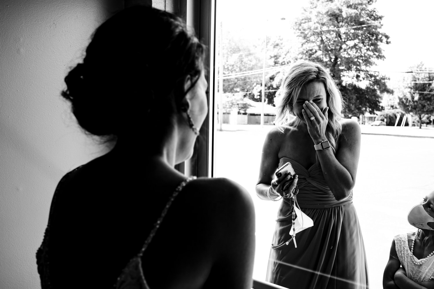 A candid black and white picture of a bride's sister in a bridesmaid gown wiping tears from her eyes as she talks to the bride through a glass door on the afternoon of a summer wedding ceremony in Kansas City. 