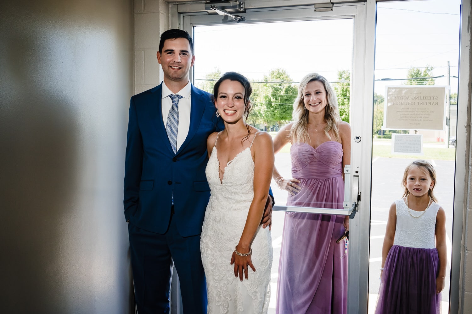 A posed picture of a bride and groom inside a church, and a bridesmaid and flower girl standing outside a glass door behind them on the afternoon of an intimate church ceremony wedding in Kansas City. 