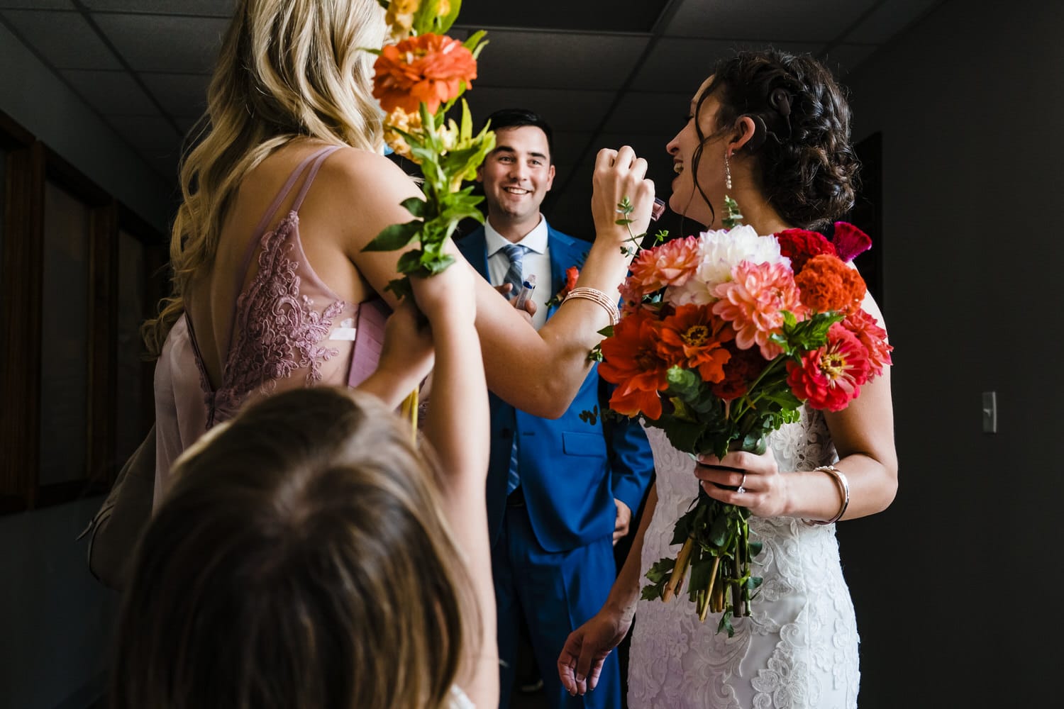 A colorful, candid picture of a bridesmaid applying lip gloss to a bride holding a bouquet as her groom looks on, laughing in the background. 