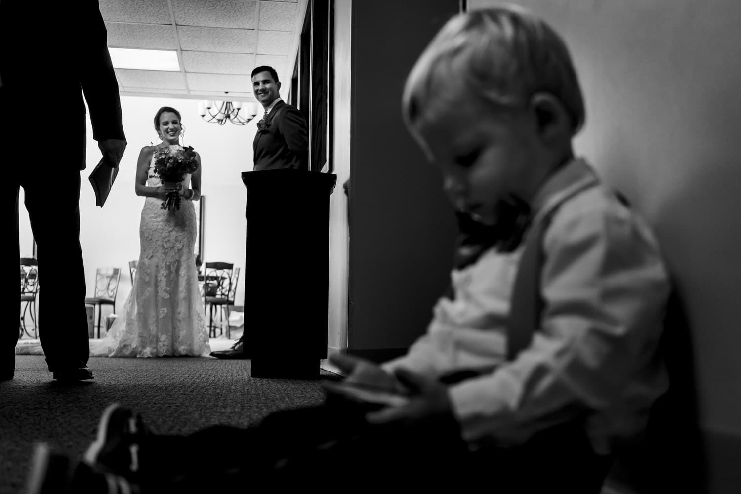 A candid black and white picture of a tiny ring bearer watching a tv show on an iPad as a bride and groom watch him and laugh moments before they walk down the aisle. 