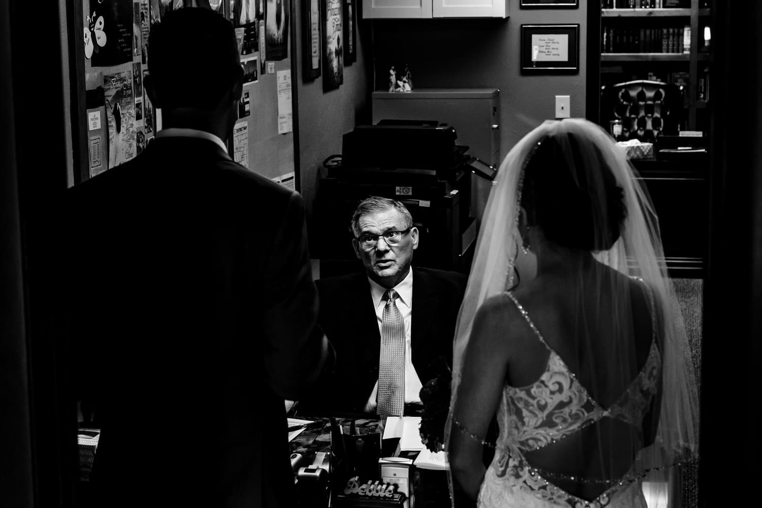 A candid black and white picture taken over the shoulders of the bride and groom as they talk to their wedding officiate. 
