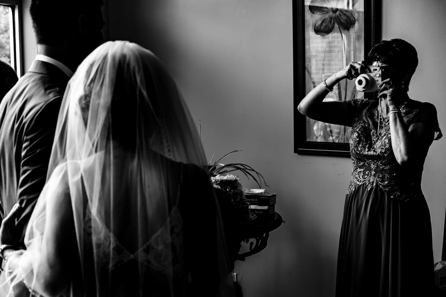 A candid black and white picture of a groom's mom taking a picture of a bride and groom with a polaroid camera on the day of an intimate church wedding ceremony in Kansas City. 