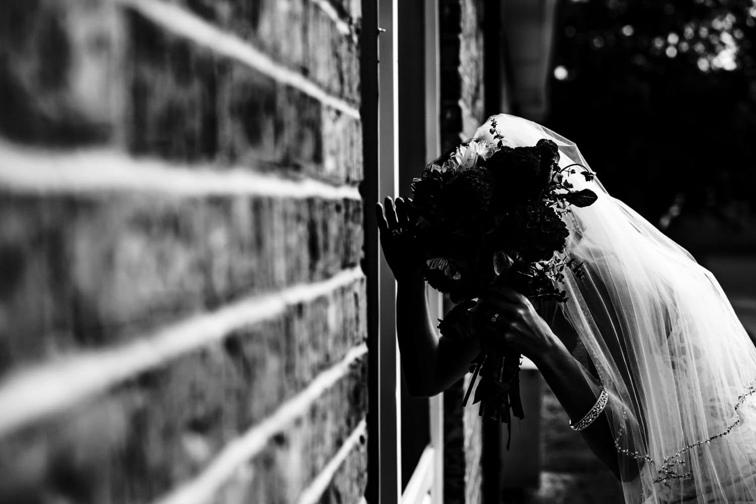 A candid black and white picture of a bride peeking into a window of a building. 