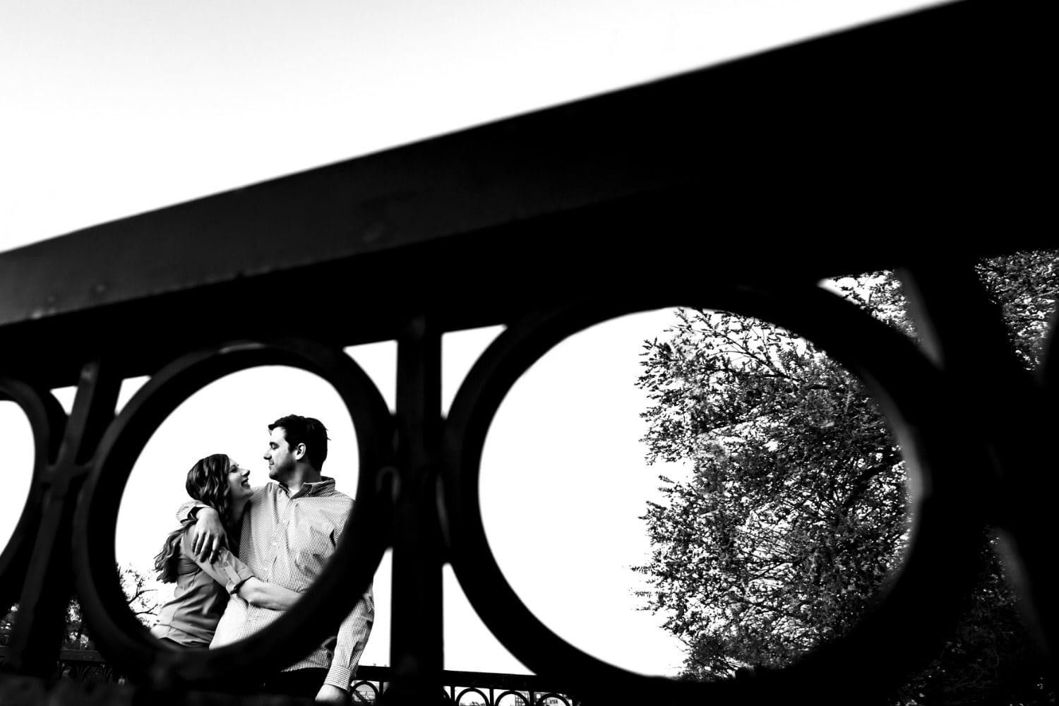 A candid black and white picture taken through a round fencing of an engaged couple holding on to each other and leaning in to share a kiss during their spring engagement session on The Plaza in Kansas City. 