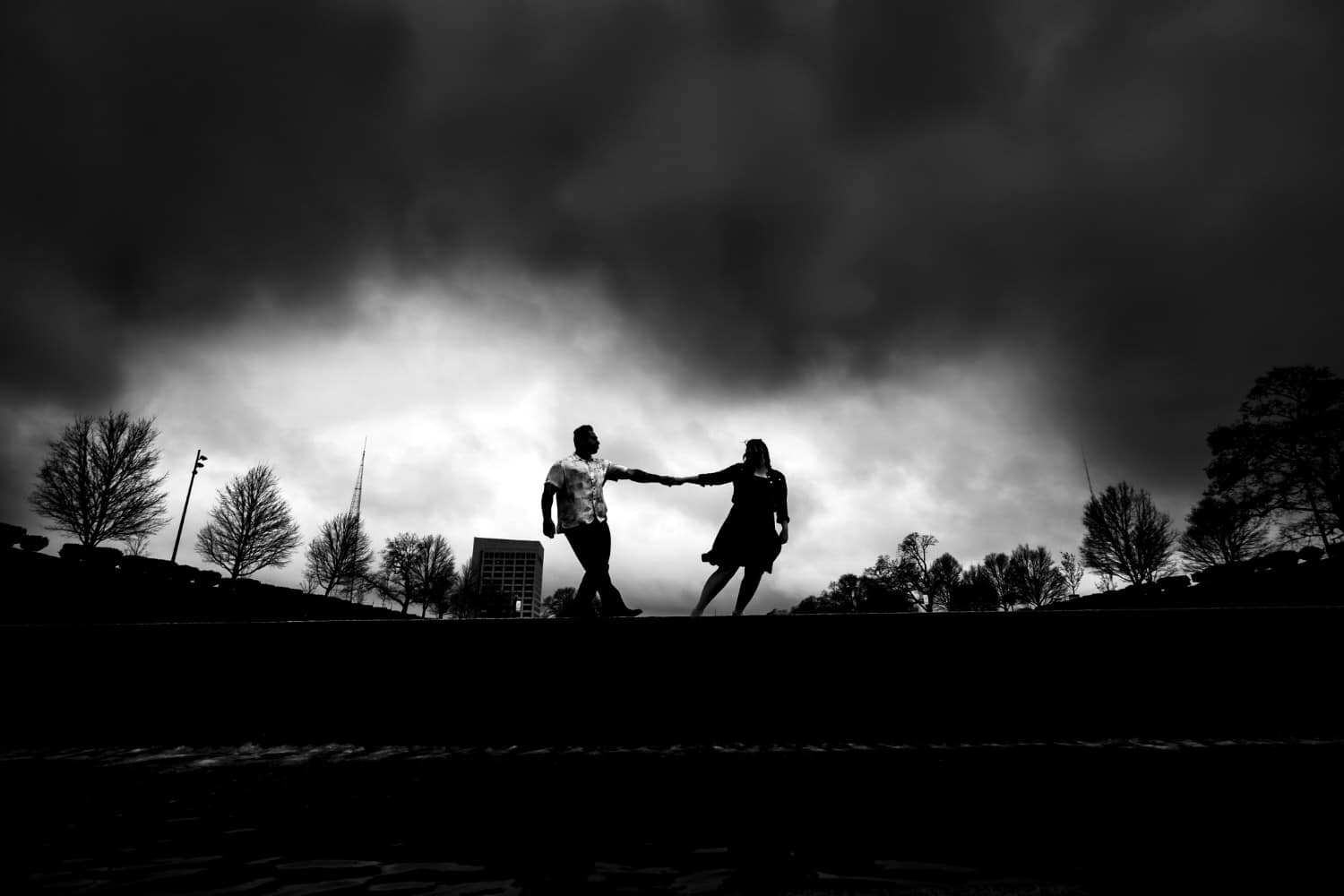 A dramatic black and white picture of a woman holding a man's hand, leading him across the horizon against a backdrop of cloudy, stormy skies during their moody engagement session in Kansas City at Liberty Memorial. 