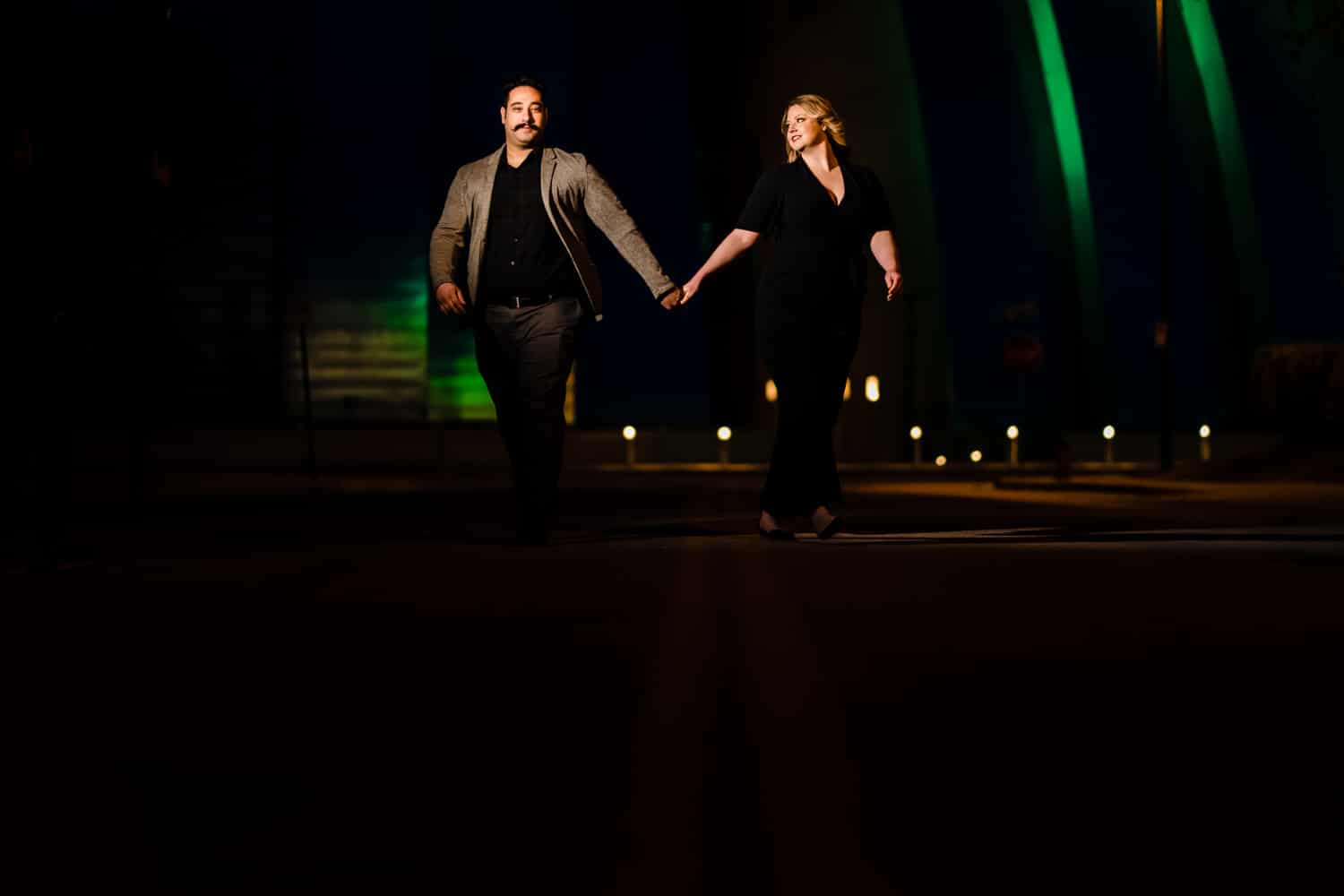 A colorful, candid portrait of an engaged couple holding hands as they walk down the center of a city street at night, the Kauffman Center for the Performing Arts visible behind them during their nighttime downtown KC engagement session. 