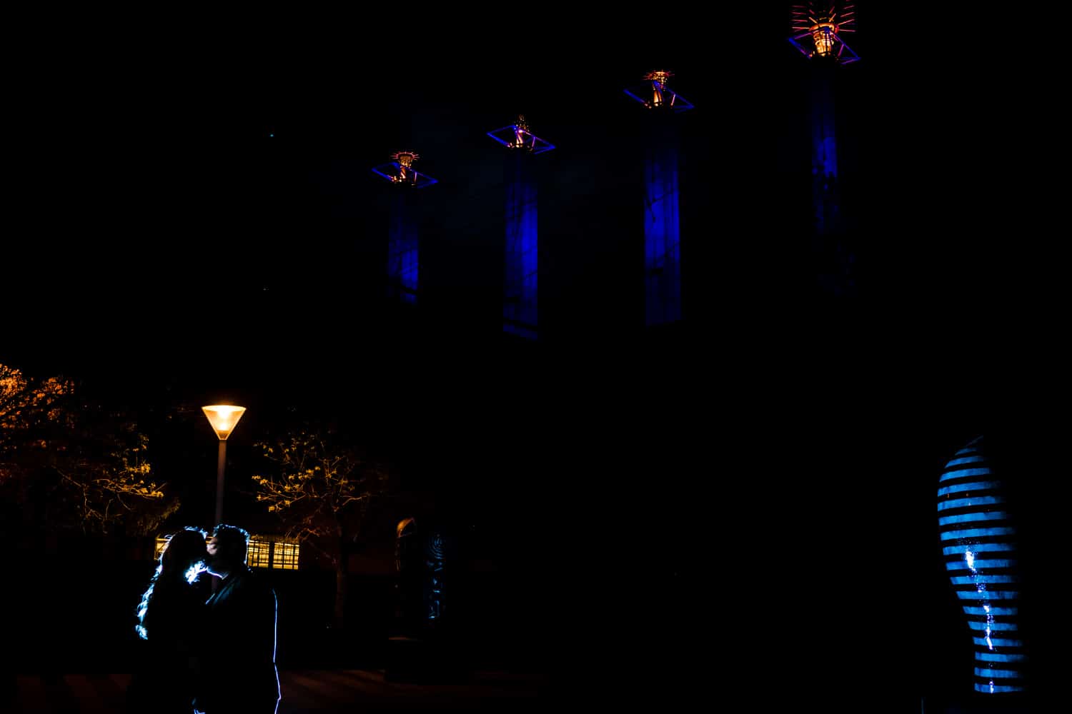 A colorful, silhouetted image of an engaged couple sharing a kiss with the infamous Kansas City spires visible behind them during their nighttime downtown KC engagement session. 