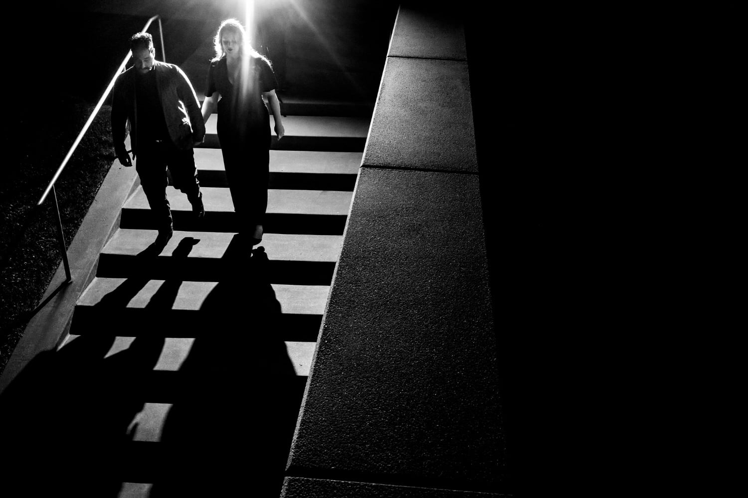 A candid, black and white picture of an engaged couple holding hands as they walk down a set of stairs, their shadows visible on the stairs in front of them during their nighttime downtown Kansas City engagement session. 