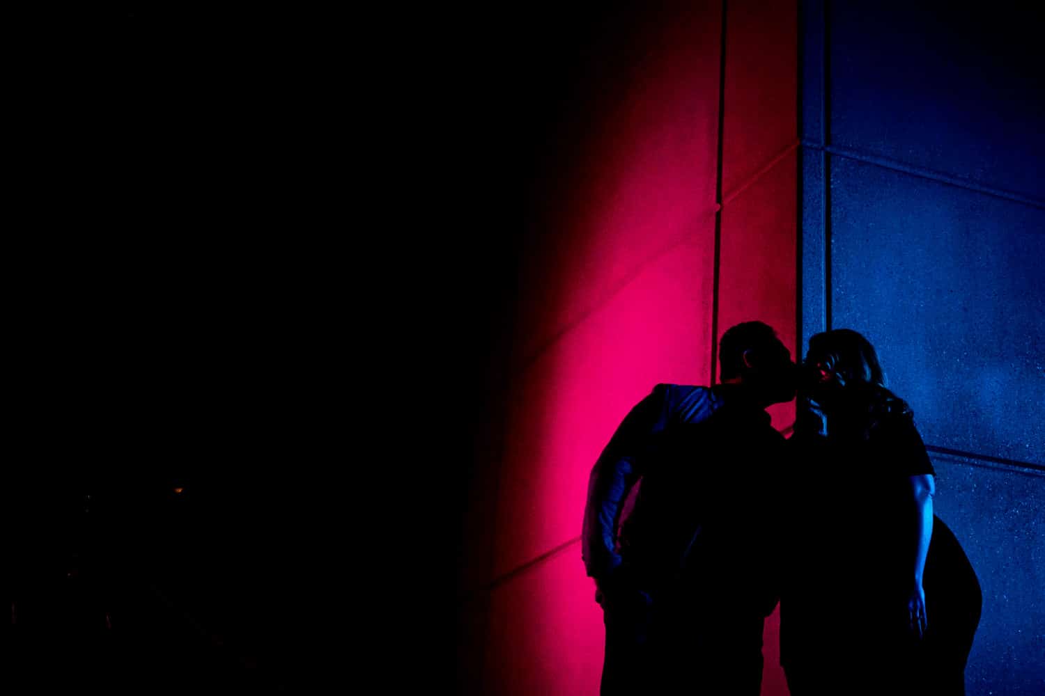 The silhouettes of an engaged couple learning in to share a kiss as they stand against a wall during their nighttime downtown Kansas City engagement session. 