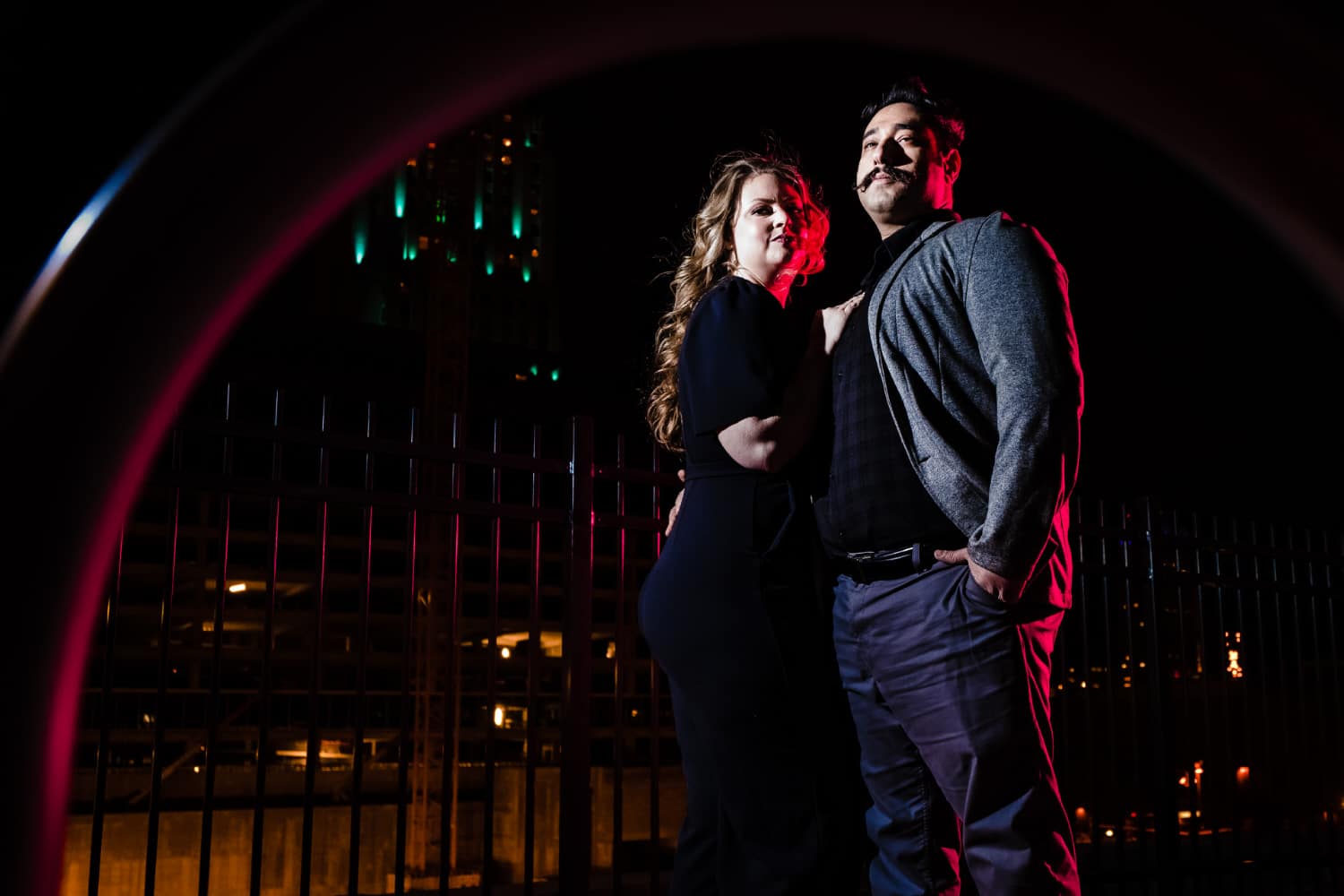 A posed portrait of an engaged couple standing side by side, the Kansas City skyline visible behind them during their nighttime engagement session. 