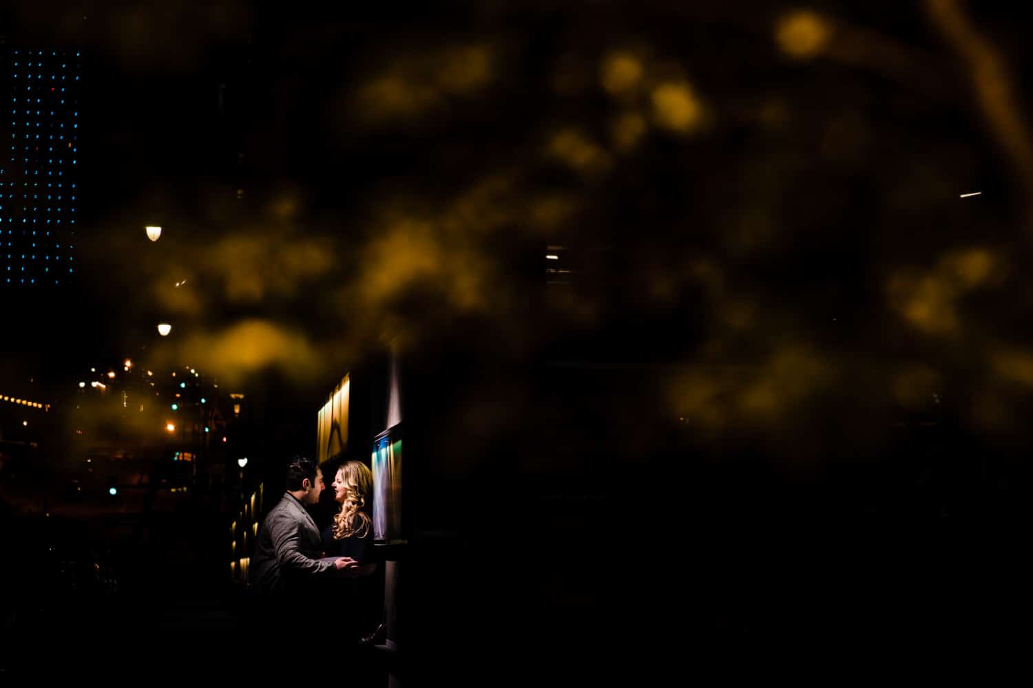 A wide, candid picture of an engaged couple leaning in to share a kiss during their nighttime downtown Kansas City engagement session. 