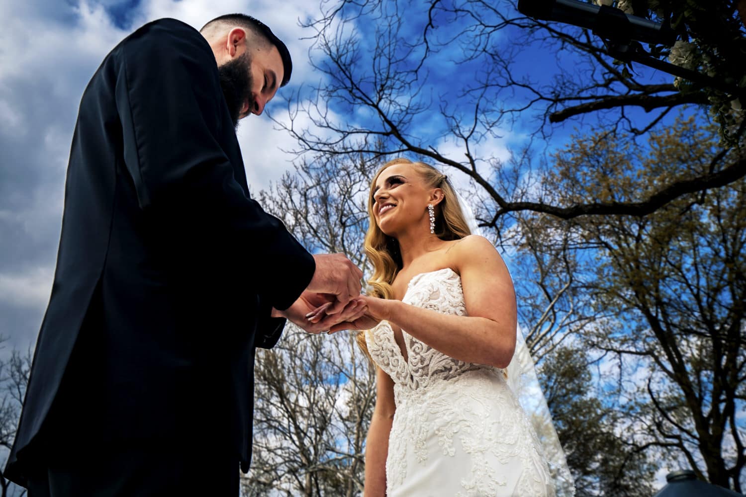 A candid picture taken during a spring wedding ceremony at the Farmhouse KC Event Venue of a groom putting a wedding band on his bride's finger, taken from the ground looking up against a bright blue sky. 