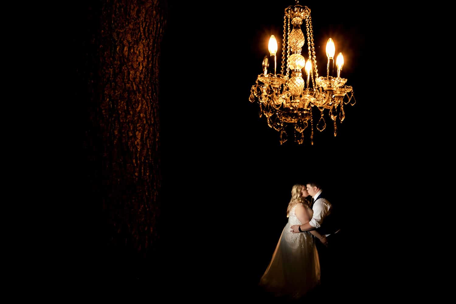 A dramatic portrait of a bride and groom sharing a hug and kiss as they stand underneath a glowing chandelier hung from a tree on the night of their summer wedding day at The Farmhouse KC Event Venue. 
