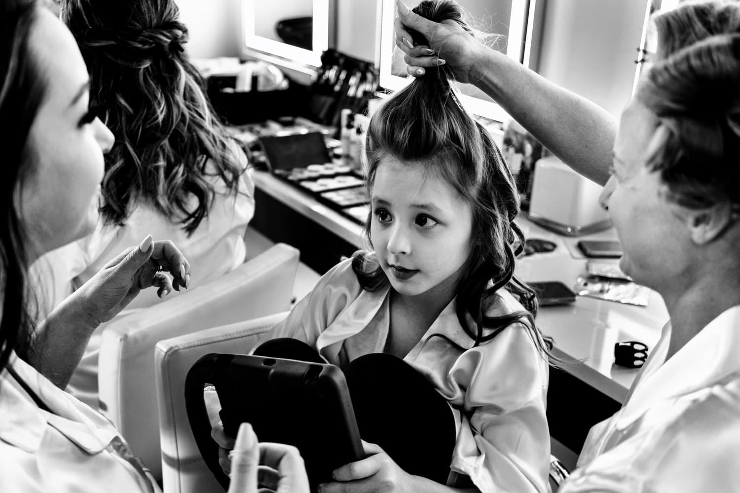 A candid black and white picture of a woman in curlers holding a little girl's hair up above her head in a ponytail in the bridal suite of The Farmhouse KC Event Venue on a spring wedding morning. 