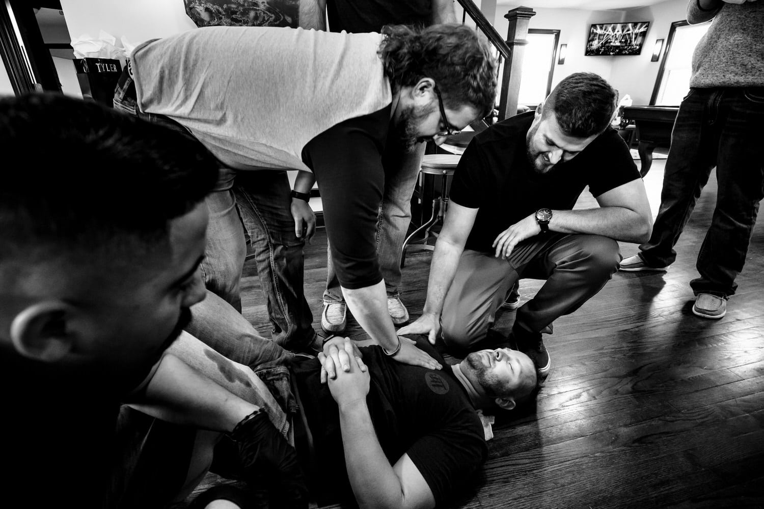 A candid black and white picture of a man lying on the floor, passed out, as his friends check on him. 