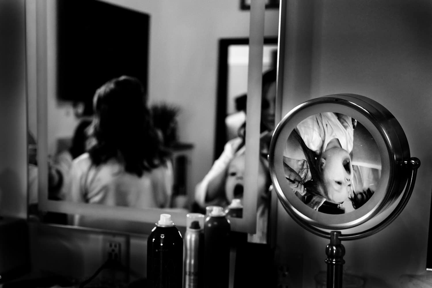 A candid black and white picture of a vanity mirror, the upside down reflection of a little girl with her mouth open visible in the reflection in the bridal suite at The Farmhouse KC Event Venue on the morning of a spring wedding. 