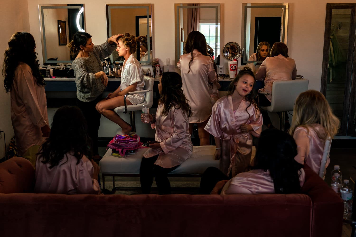 A wide, colorful, candid picture of the full bridal suite at The Farmhouse KC Event Venue as a bride gets her makeup done on her wedding morning. 