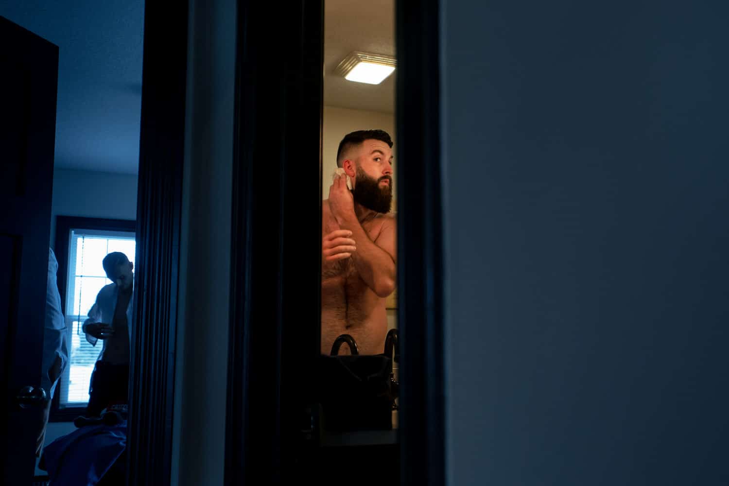 A candid picture of a man without a shirt on touching up his beard in a bathroom on the morning of his spring wedding. 