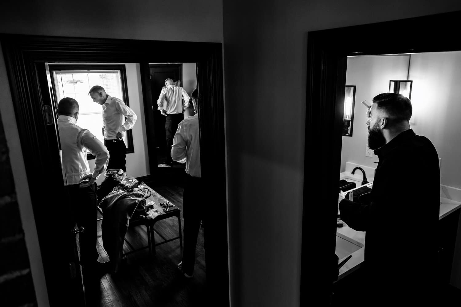 A wide, black and white picture of a man in a bathroom fixing his hair as his friends get dressed in the room next door on the morning of a spring wedding in Kansas City. 