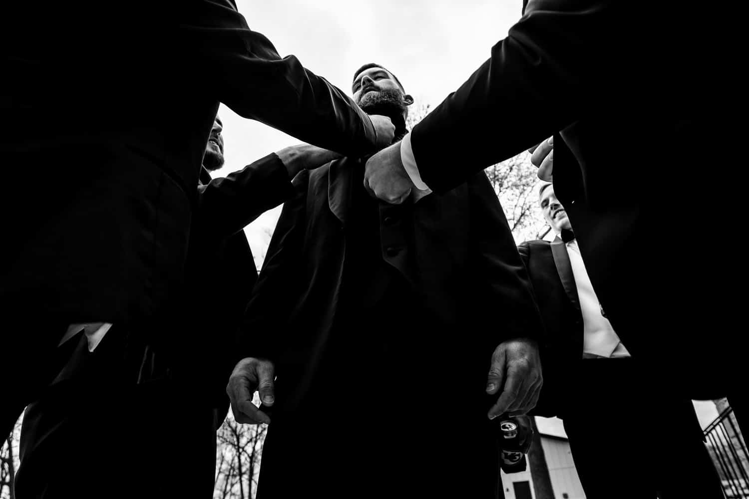 A candid black and white picture taken from the ground looking up of a groom's friends fixing his tie and jacket on the morning of his wedding. 