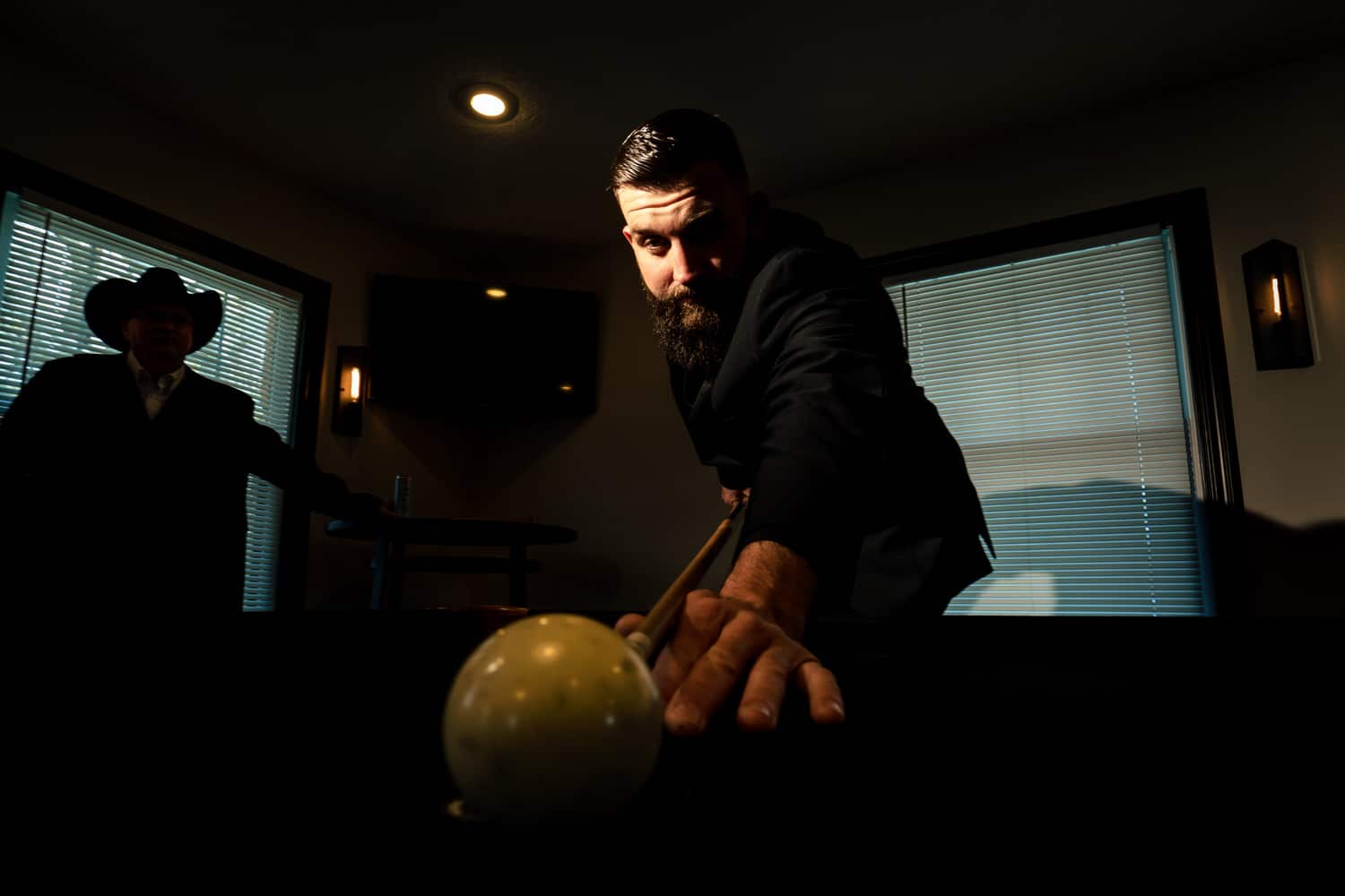 A picture of a man in a black tuxedo bending over a pool table to hit a cue ball. 
