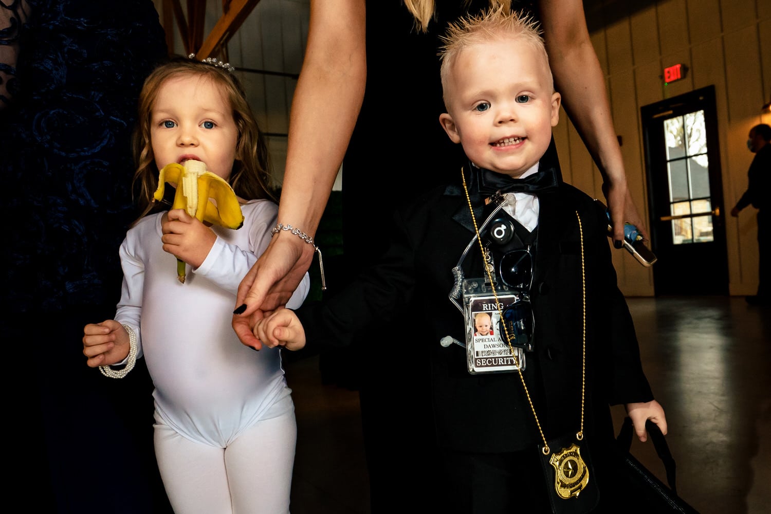 A candid picture of a ring bearer with a badge and a flower girl in a leotard eating a banana. 
